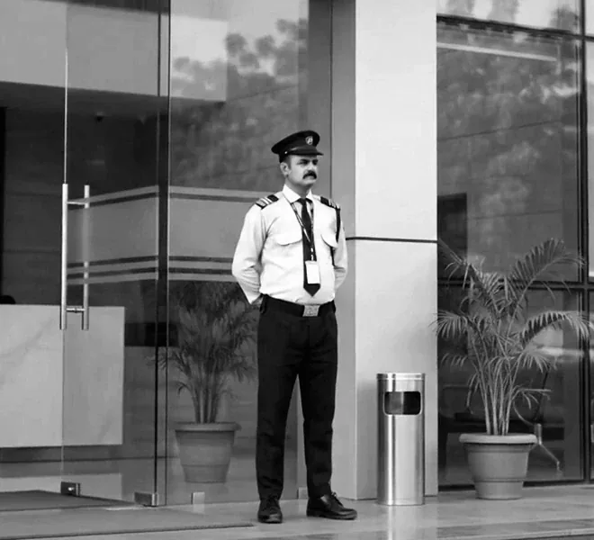 A security officer in uniform standing at the glass doors of an office building